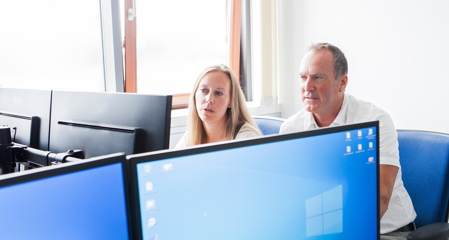 Zwei Personen, eine Frau und ein Mann, sitzen gemeinsam vor mehreren Bildschirmen in einem hellen Büro mit Fenster. Sie konzentrieren sich auf die Monitore, was eine Beratung oder Zusammenarbeit im Bereich IT-Support oder IT-Beratung nahelegt.