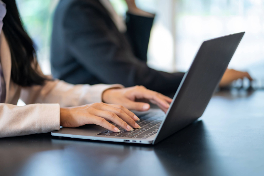 Hände einer Frau tippen auf Laptop-Tastatur, im Hintergrund unscharf weitere Person am Computer, symbolisiert IT-Beratung, IT-Support und digitale Kommunikation in einem modernen Arbeitsumfeld.