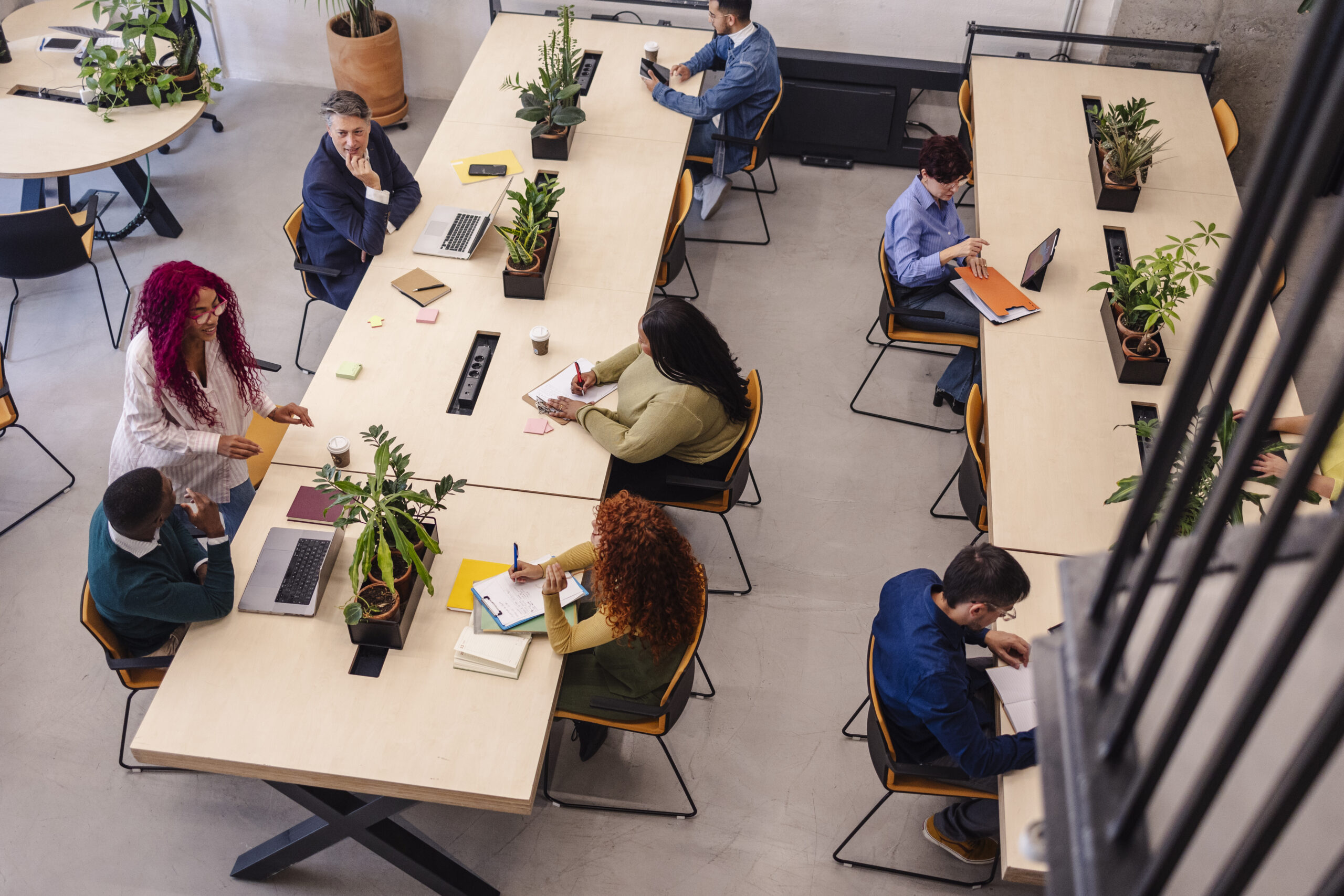 Mehrere Mitarbeitende arbeiten in einem modernen, hellen Büro an langen Tischen mit Laptops, Notizbüchern und Kaffeetassen. Pflanzen stehen als Dekoration auf den Tischen. Die Personen sind in Gespräche vertieft oder konzentrieren sich auf ihre Aufgaben, was eine kollaborative Arbeitsatmosphäre vermittelt.