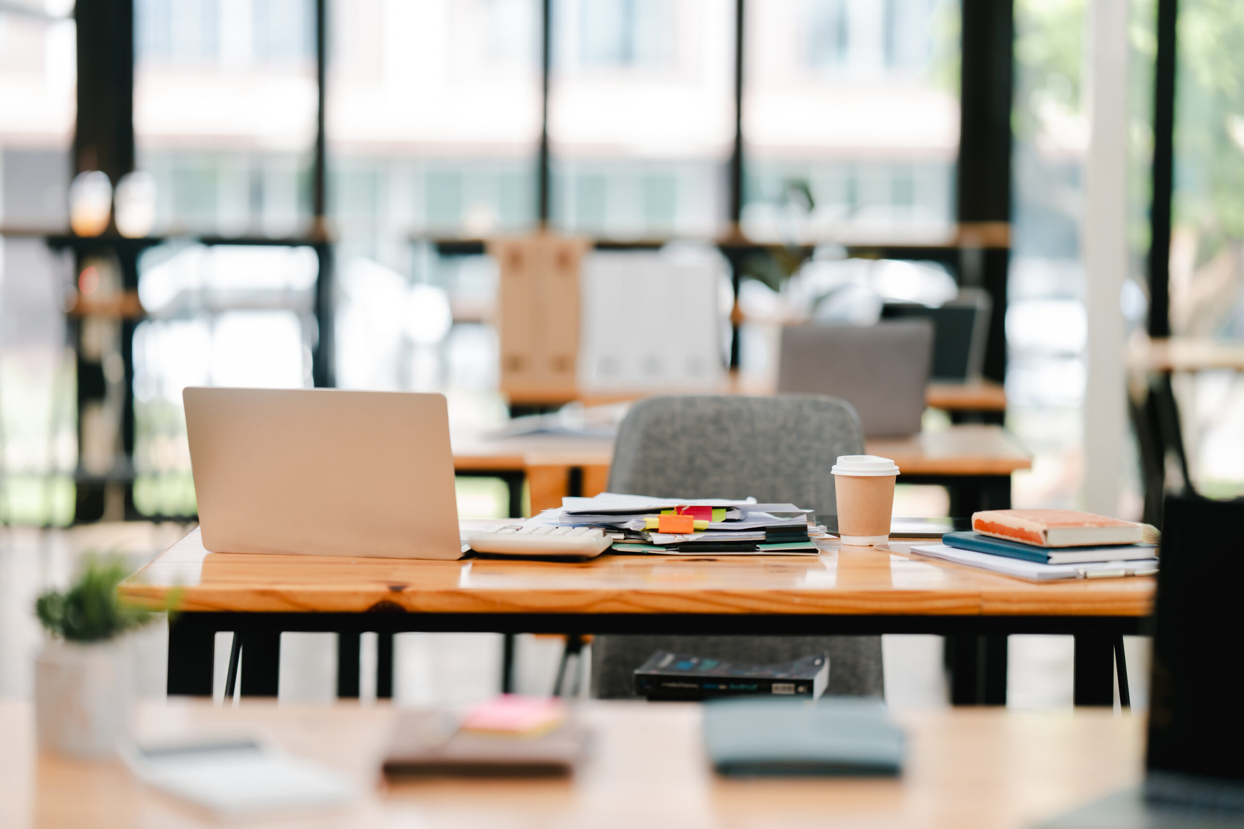 Moderner Arbeitsplatz mit Laptop, Papierstapel, Kaffeebecher und Büromaterial auf hellem Holztisch in hellem, offenem Büro mit großen Fenstern, symbolisiert IT-Systemhaus und Digitalisierung.