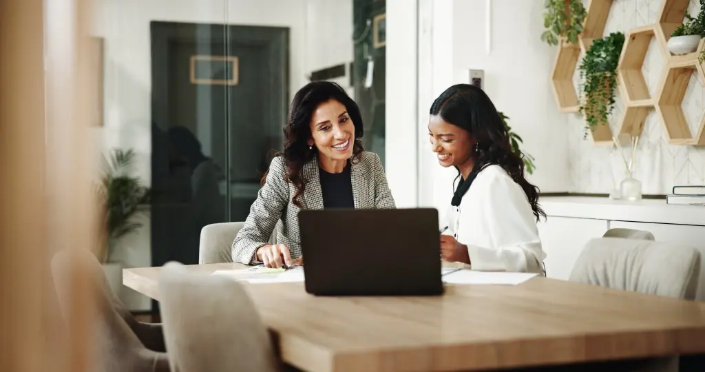 Zwei Frauen sitzen an einem Holztisch in einem modernen, hellen Büro und schauen gemeinsam auf einen Laptop, dabei lächeln sie und wirken engagiert in einem Beratungsgespräch. Im Hintergrund sind Pflanzen und dekorative Wandregale mit sechseckigen Fächern zu sehen.