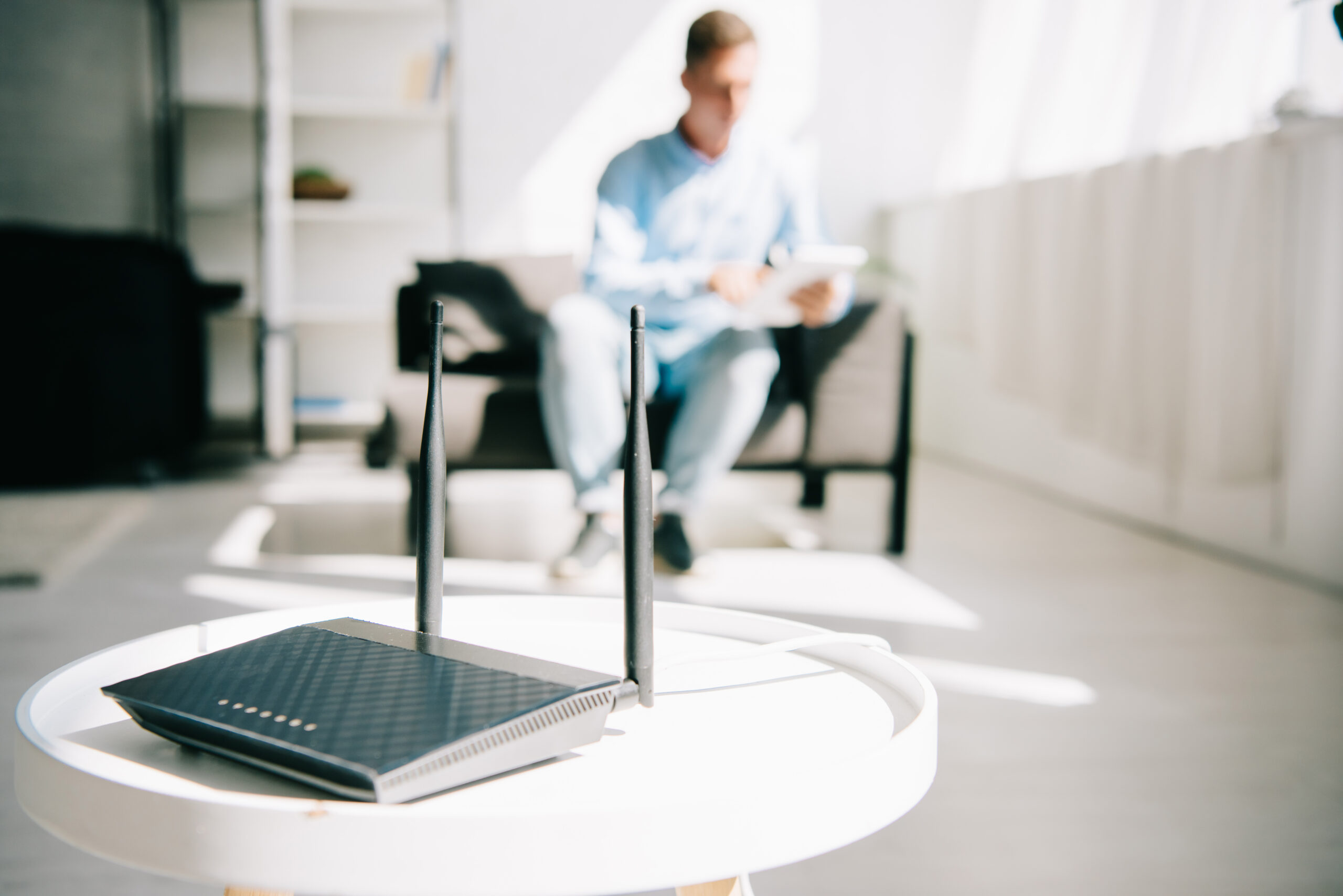 Schwarzer WLAN-Router mit zwei Antennen steht auf weißem runden Tisch in hellem Raum, im Hintergrund unscharf sitzender Mann mit Tablet auf Sofa, symbolisiert IT- und Netzwerklösungen.