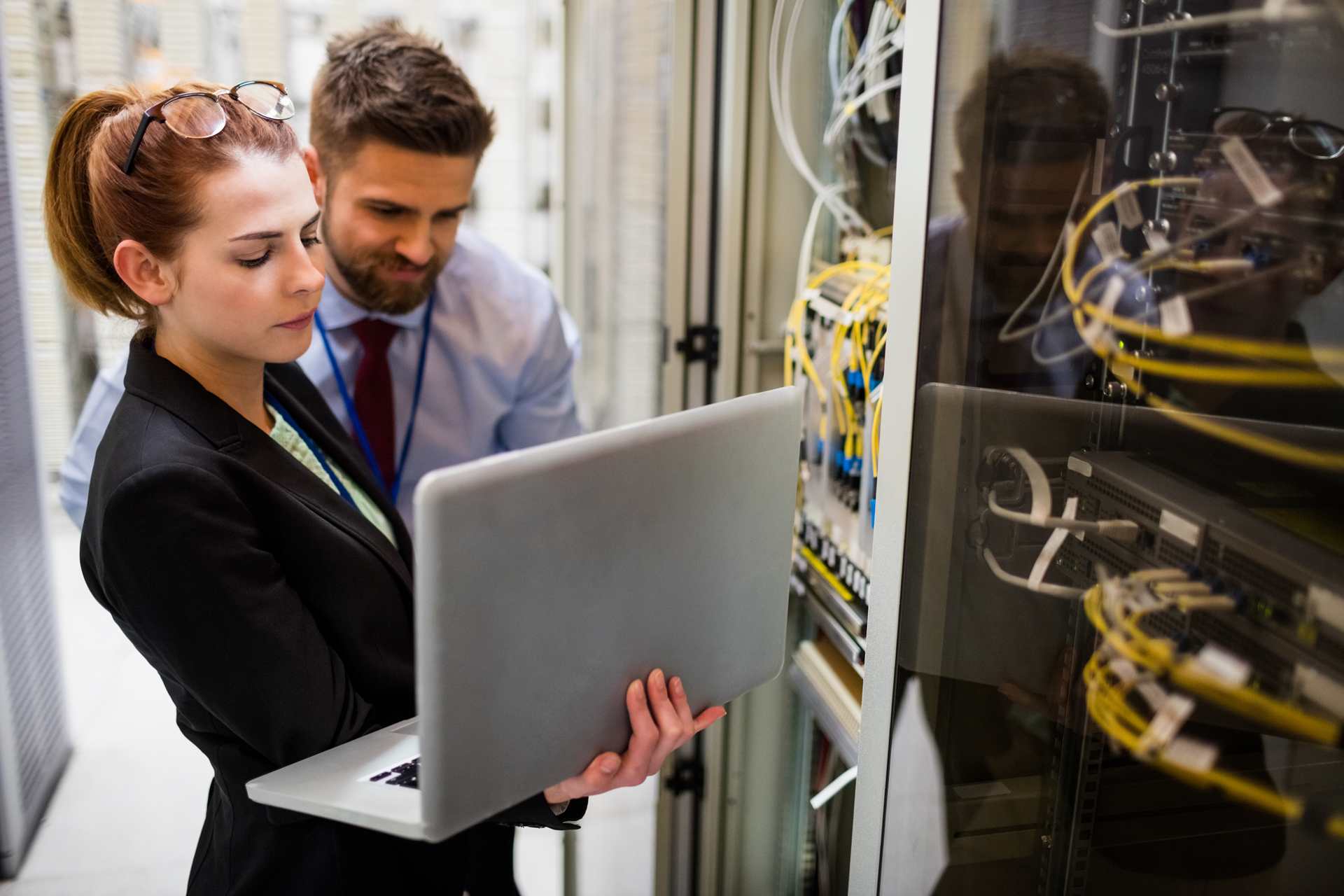Zwei Techniker, eine Frau und ein Mann, analysieren gemeinsam Daten auf einem Laptop vor einem Serverrack mit zahlreichen vernetzten Kabeln in einem Rechenzentrum, symbolisiert IT-Support und Netzwerklösungen der dierckgroup.