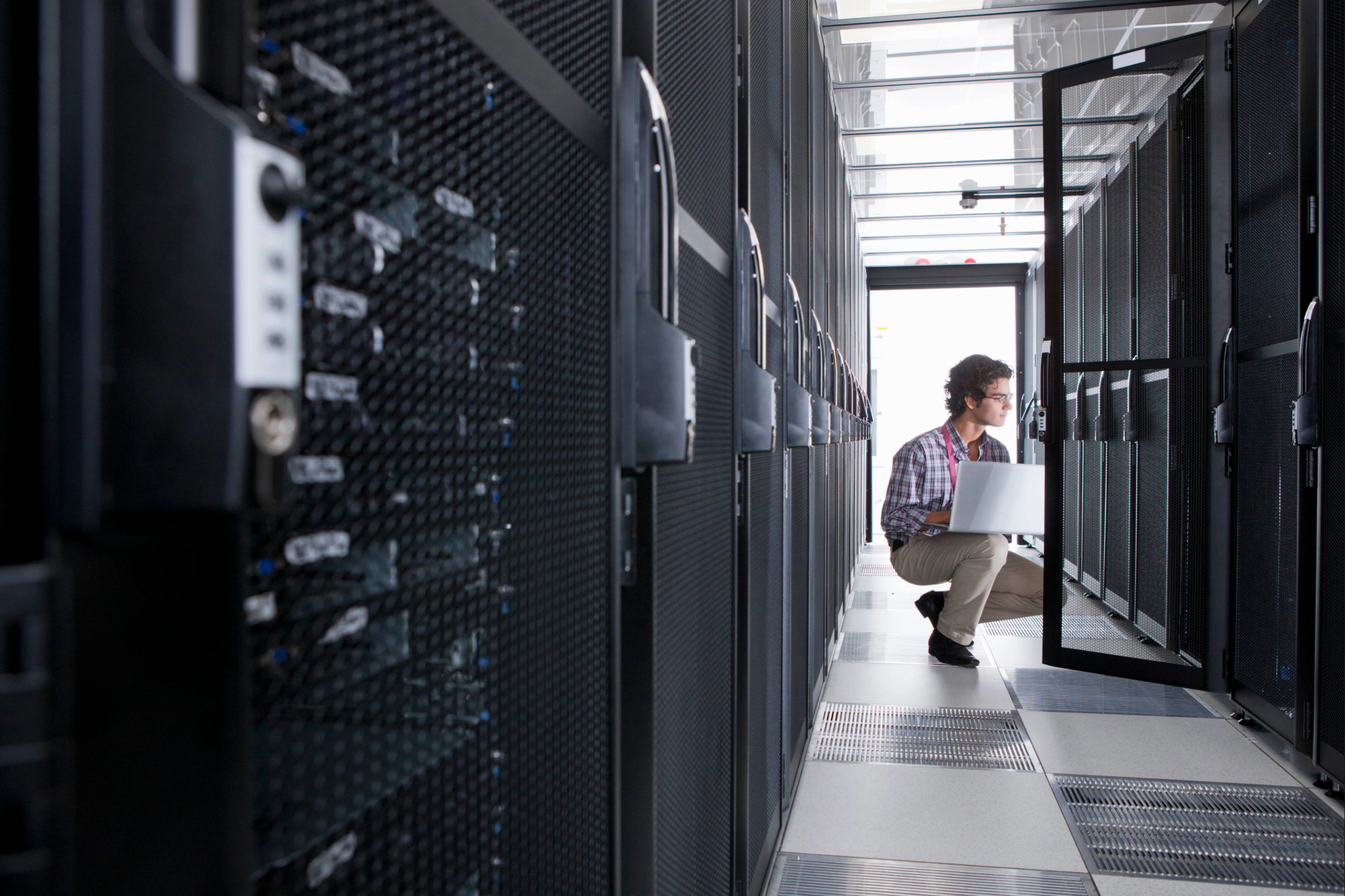 Techniker mit Laptop kniet in einem Rechenzentrum zwischen Reihen von Server-Racks, prüft IT-Systeme und Netzwerklösungen in einem modernen, gut beleuchteten Raum.