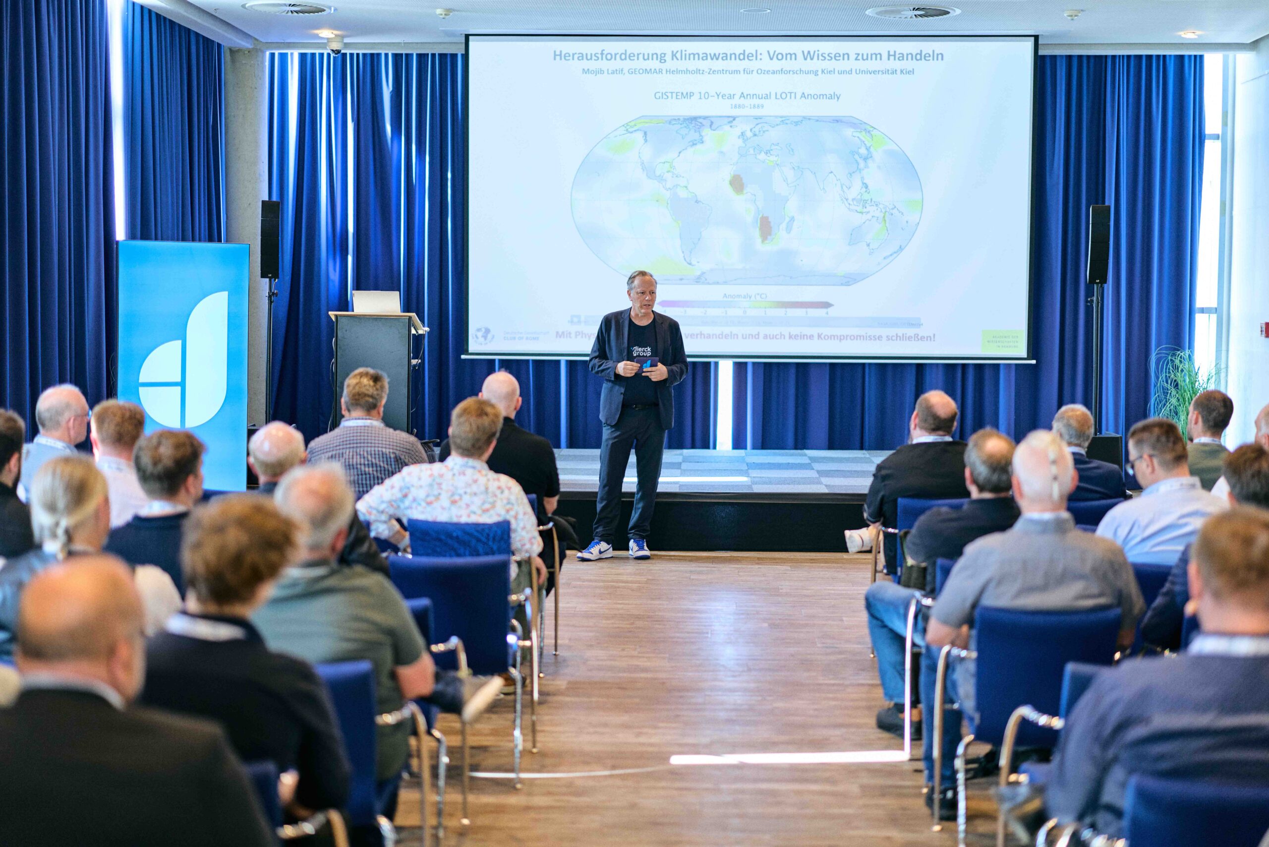 Mann hält Vortrag vor Publikum in Konferenzraum mit blauen Vorhängen, auf Leinwand Weltkarte zur Klimawandel-Herausforderung mit Text „Vom Wissen zum Handeln“, Publikum sitzt auf blauen Stühlen, links blaues Banner mit stilisiertem weißem Logo, helle Holzbodenfläche.