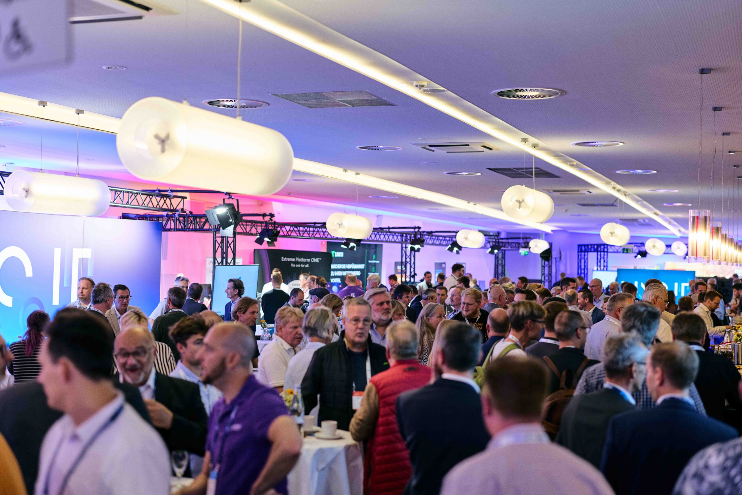 Großer, heller Konferenzraum mit zahlreichen Menschen in Business-Kleidung, die sich an Stehtischen und in Gruppen unterhalten. Moderne Deckenbeleuchtung und bunte LED-Beleuchtung an den Wänden schaffen eine professionelle Atmosphäre. Im Hintergrund sind Messestände und Bildschirme mit Präsentationen zu sehen. Veranstaltung mit Fokus auf IT, Digitalisierung und Kommunikationstechnik.