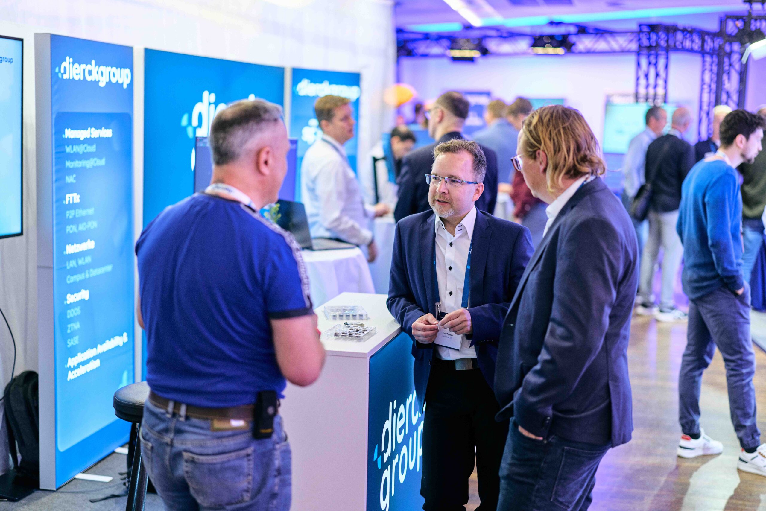 Mehrere Personen im Gespräch an einem Messestand der dierckgroup mit blauem Branding und Displays, die Managed Services und IT-Lösungen zeigen, in einer hell beleuchteten Messehalle mit weiteren Ständen und Besuchern im Hintergrund.