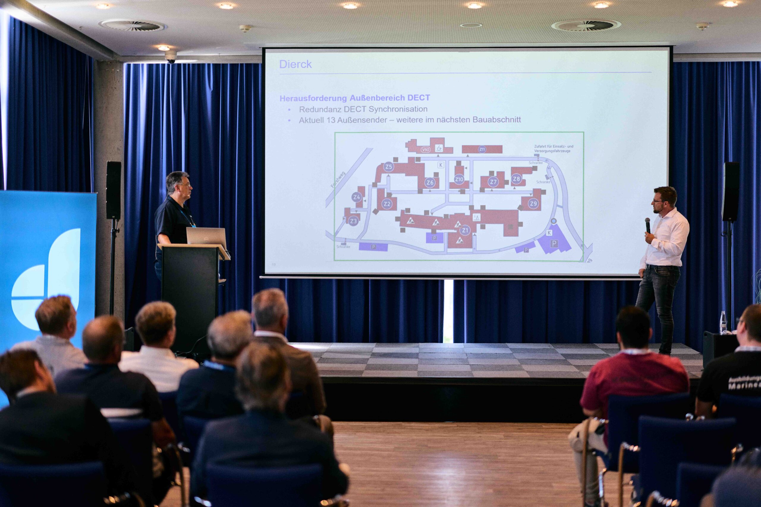 Zwei Männer präsentieren vor Publikum in einem Konferenzraum eine Folie mit dem Titel „Herausforderung Außenbereich DECT“ und einer schematischen Karte eines Gebäudekomplexes. Im Hintergrund dunkelblaue Vorhänge, links ein blaues Banner mit weißem Logo. Die Szene vermittelt eine professionelle IT-Beratung oder Fachvortrag.