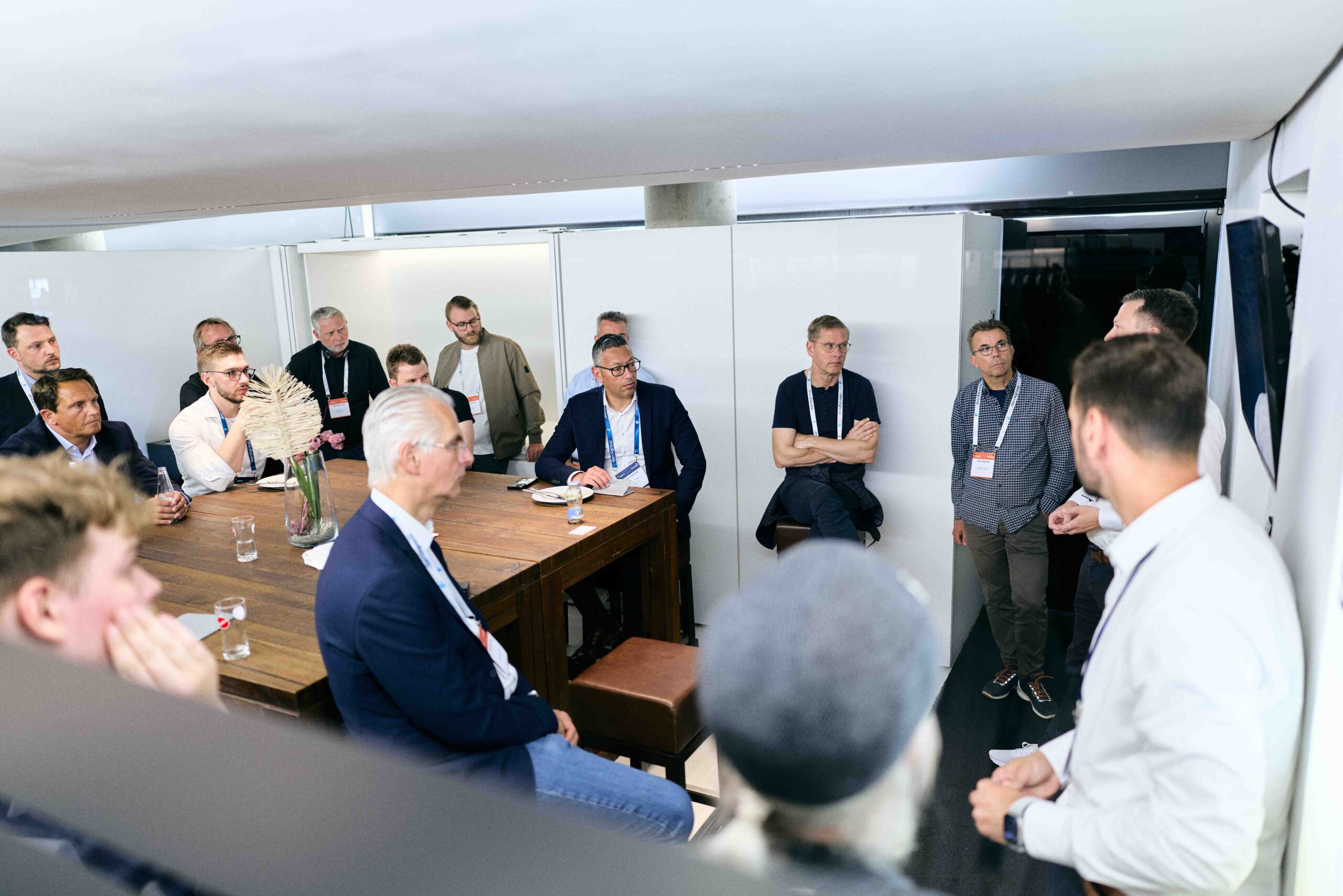 Mehrere Männer unterschiedlichen Alters sitzen und stehen in einem modernen, hellen Besprechungsraum um einen großen Holztisch, hören aufmerksam einem stehenden Sprecher zu, der eine Präsentation hält. Die Atmosphäre wirkt professionell und konzentriert.