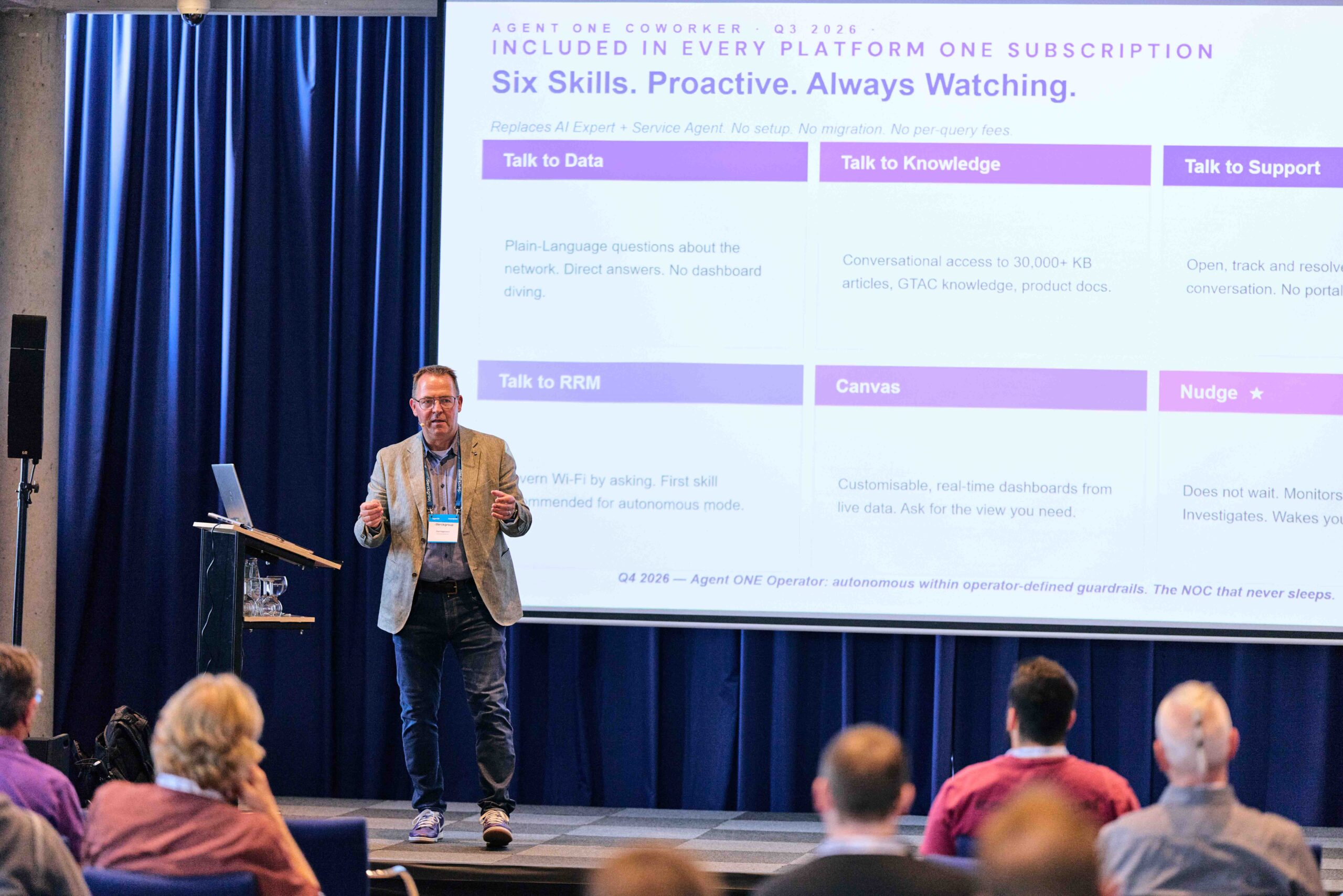 Mann in grauem Sakko und Jeans hält Präsentation vor Publikum in Konferenzraum mit blauem Vorhang, auf großer Leinwand sind Stichpunkte zu Plattform-Funktionen wie „Talk to Data“, „Talk to Knowledge“ und „Talk to Support“ zu sehen, Szene vermittelt IT-Beratung und Digitalisierung.