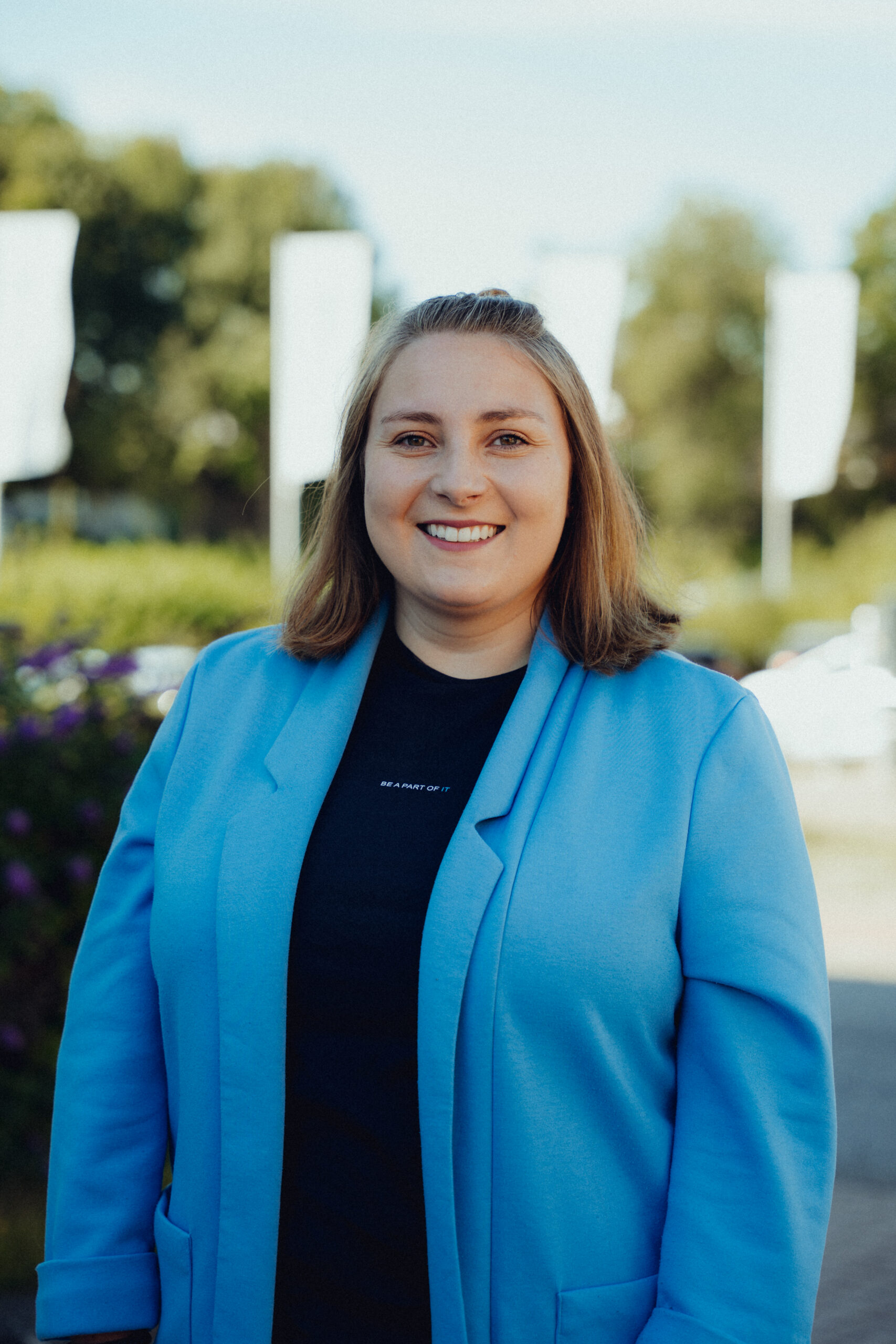 Junge Frau mit schulterlangen hellbraunen Haaren, lächelnd, trägt hellblauen Blazer über schwarzem Shirt mit kleinem weißen Schriftzug „BE A PART OF IT“, vor unscharfem Hintergrund mit Bäumen und weißen Fahnen, symbolisiert freundlichen Kundenkontakt und IT-Beratung bei dierckgroup.