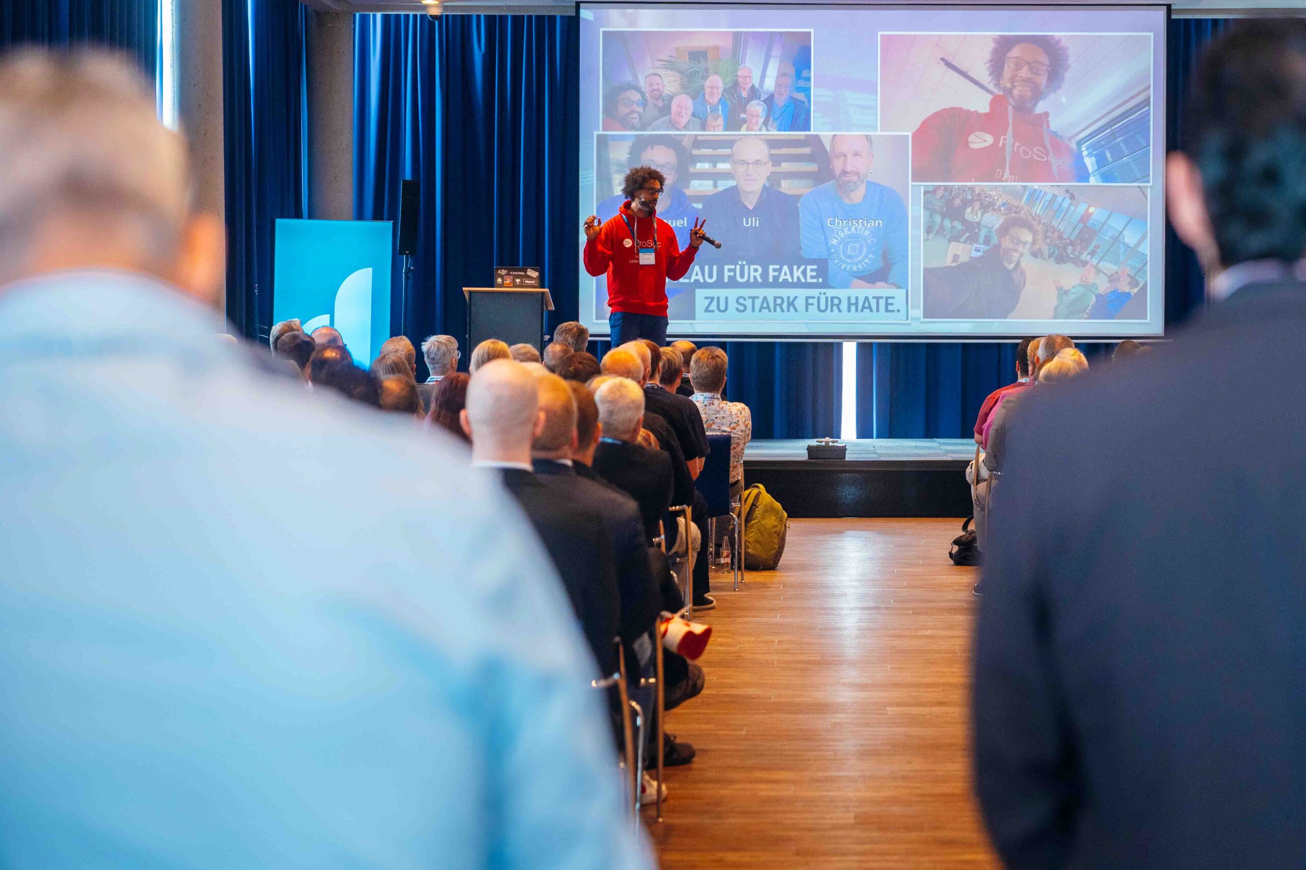 Vortragender mit rotem Pullover spricht vor Publikum in einem Konferenzraum, im Hintergrund große Leinwand mit mehreren Porträtfotos und Textfragment „...AU FÜR FAKE. ... ZU STARK FÜR HATE.“ Publikum sitzt auf Stühlen, Raum mit Holzboden und blauen Vorhängen, Veranstaltung im Kontext von IT-Lösungen und Digitalisierung.
