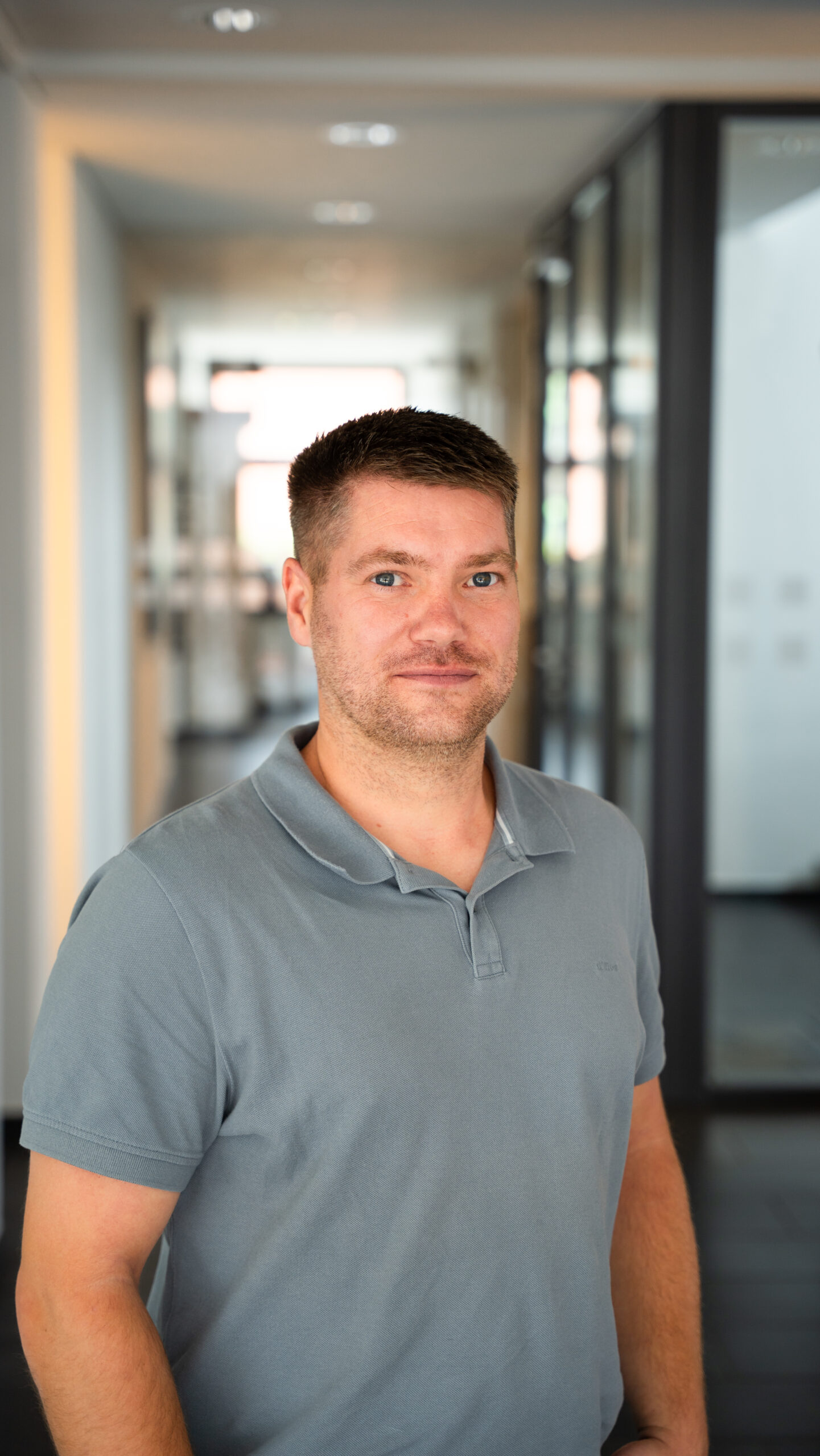 Mann mit kurzem braunem Haar und grauem Poloshirt steht in einem modernen Büroflur mit Glaswänden, neutraler Hintergrund mit weichem Licht, professionelle Atmosphäre passend zu IT-Beratung und Managed Services bei dierckgroup.