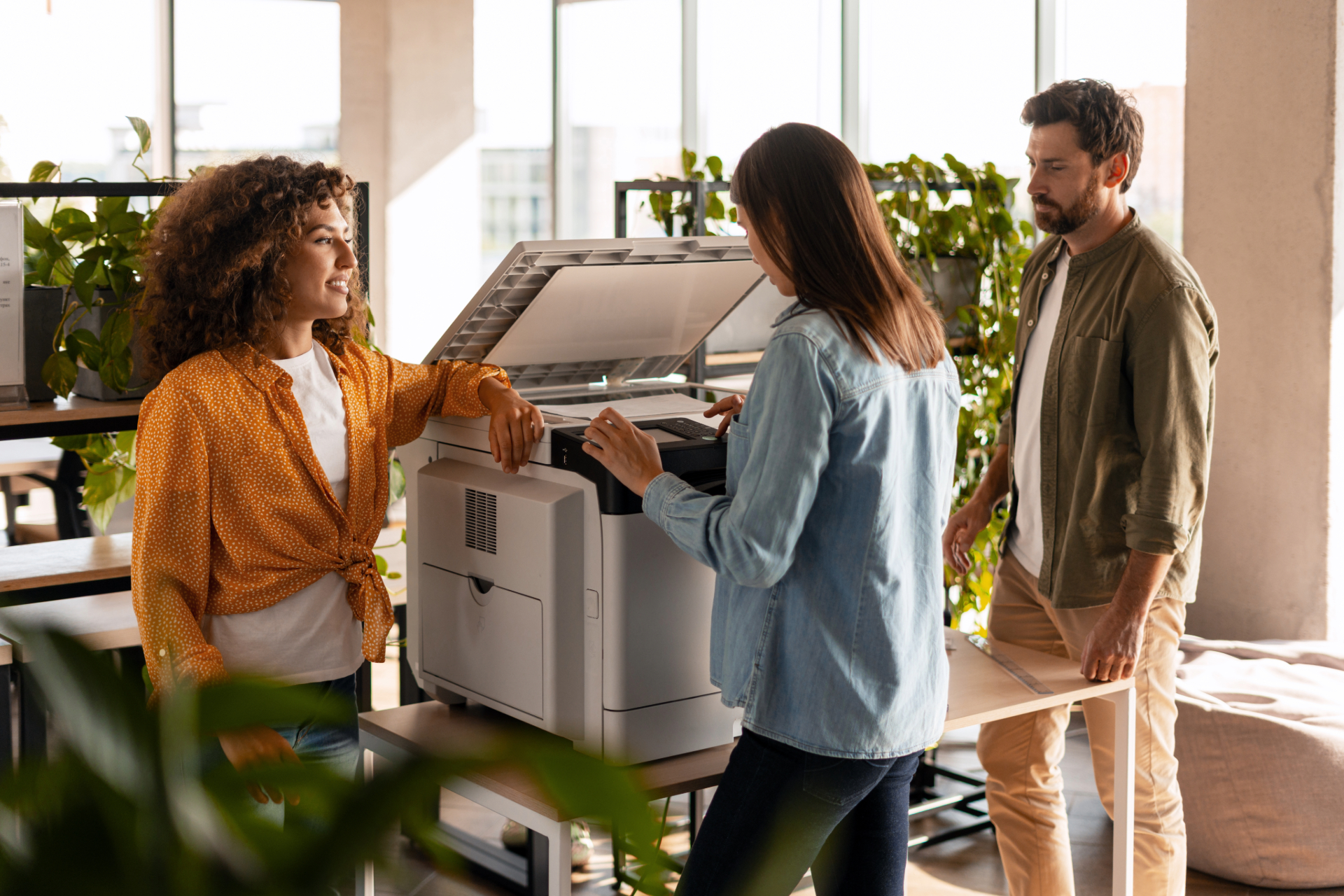Drei Personen in modernem Büro arbeiten gemeinsam an einem Multifunktionsdrucker, eine Frau bedient das Gerät, während die anderen beiden aufmerksam zuschauen. Helle, freundliche Arbeitsumgebung mit großen Fenstern und Pflanzen im Hintergrund, symbolisiert Zusammenarbeit und IT-Support im Bereich Scan and Printing.