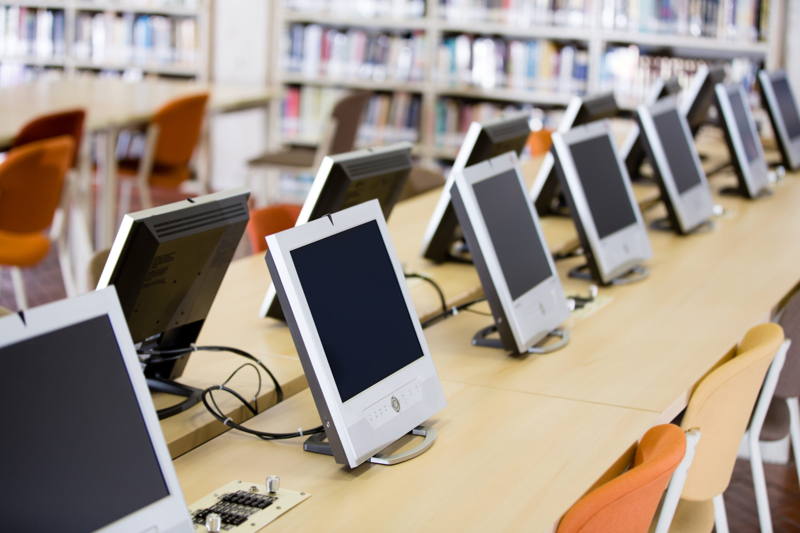 Reihe von ausgeschalteten Computermonitoren auf einem langen Holztisch in einem hellen Raum mit Bücherregalen im Hintergrund, mehrere orangefarbene Stühle an den Arbeitsplätzen, symbolisiert IT-Systemhaus und Arbeitsplatzlösungen für moderne IT-Umgebungen.