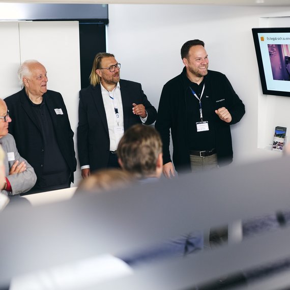 Drei Männer in Business-Kleidung stehen vor einem Bildschirm und präsentieren in einem modernen Besprechungsraum, im Vordergrund unscharfe Köpfe von Zuhörern. Der Bildschirm zeigt eine Folie mit Text und Bild, die Präsentation wirkt professionell und freundlich.