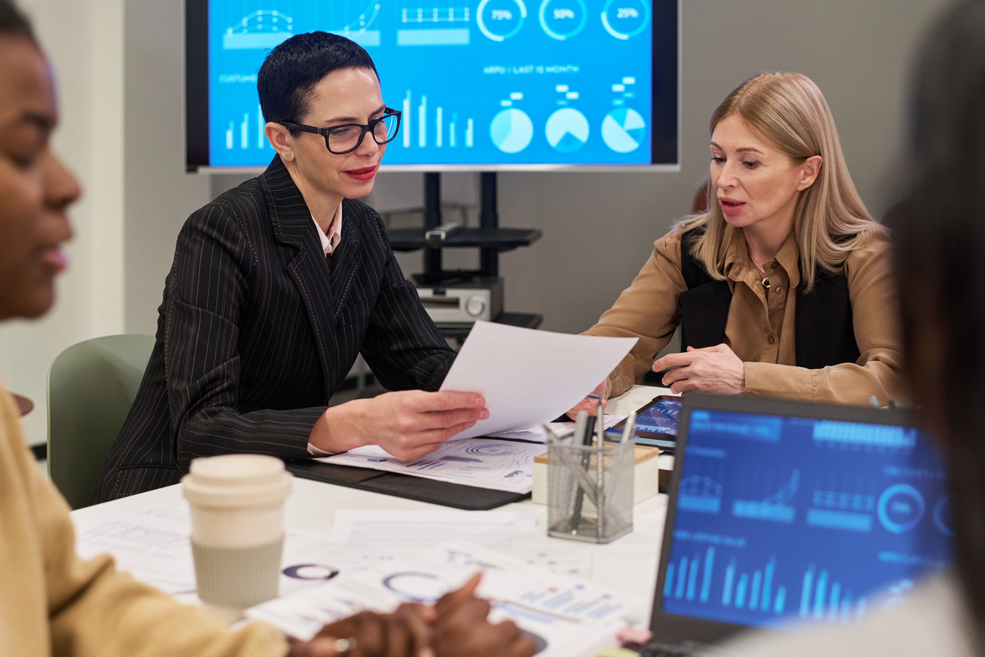 Mehrere Frauen sitzen an einem Tisch in einem modernen Büro und besprechen Geschäftsdaten. Auf dem Tisch liegen Ausdrucke mit Diagrammen und Grafiken, im Hintergrund sind Bildschirme mit weiteren Datenvisualisierungen zu sehen. Die Szene vermittelt Zusammenarbeit und Analyse, passend zu IT-Beratung, Managed Services und Digitalisierung.