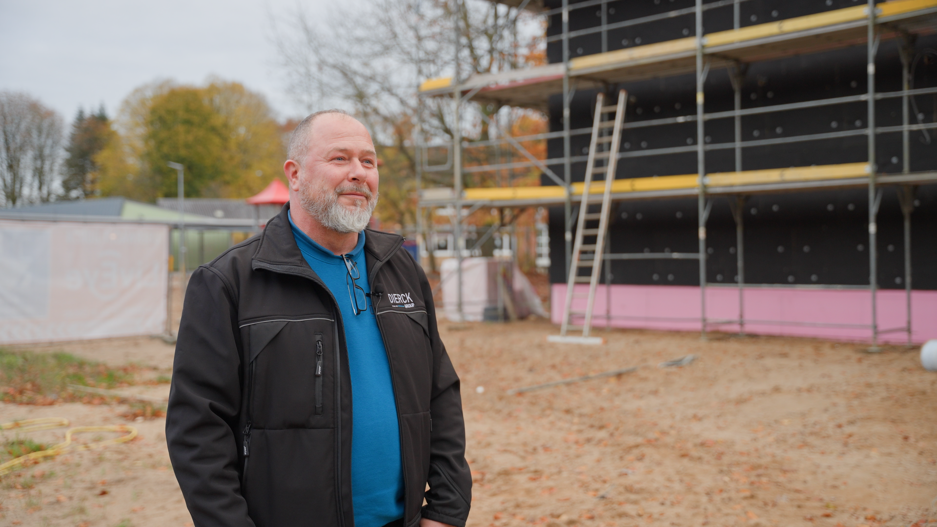 Mann mit grauem Bart und kurzem Haar trägt schwarze dierckgroup-Jacke und blaues Shirt, steht auf einer Baustelle mit Gerüst und Baugeräten im Hintergrund, herbstliche Bäume sichtbar.
