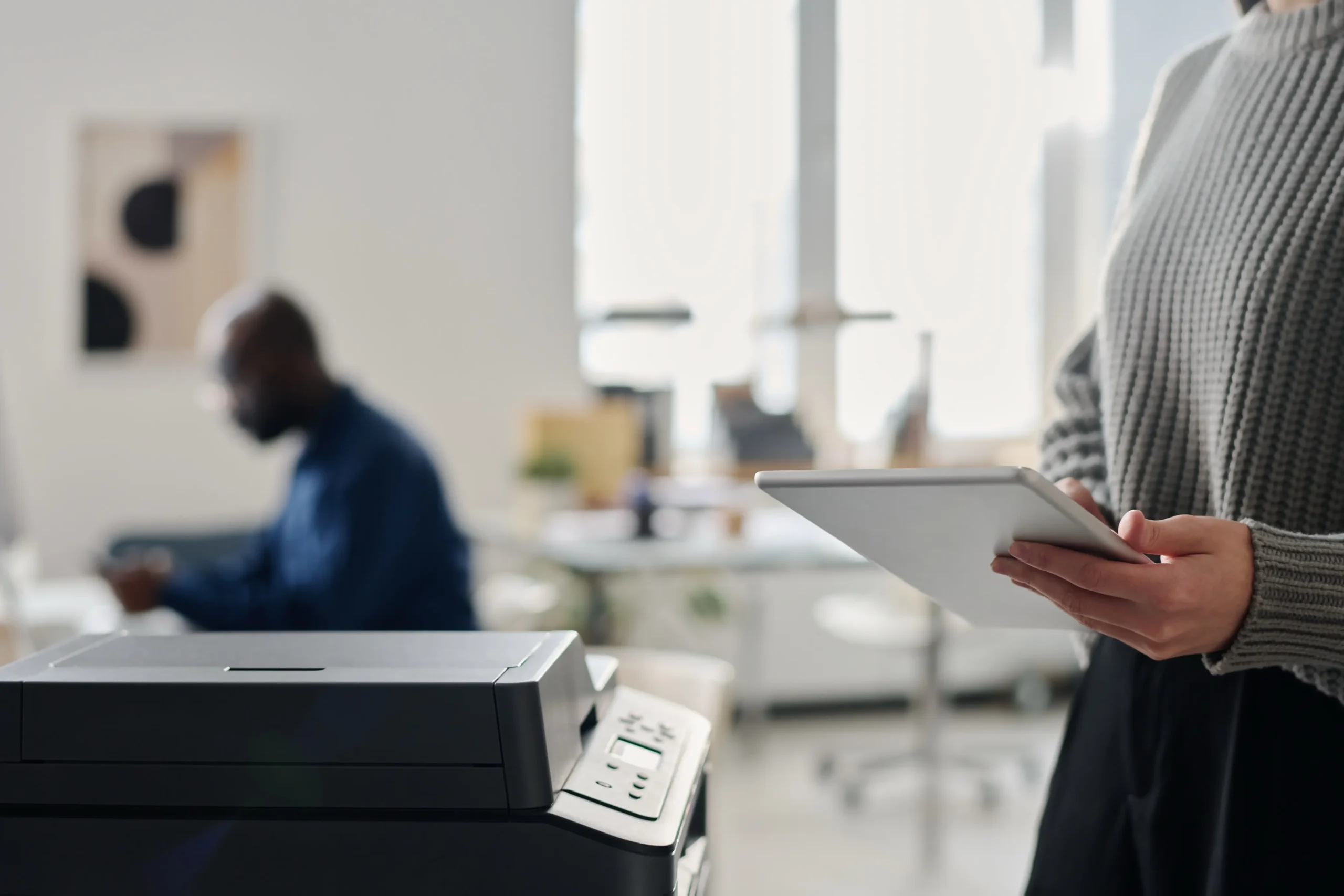 Person in grauem Pullover hält ein digitales Tablet in der Hand, im Vordergrund steht ein moderner Drucker, im Hintergrund unscharf ein weiterer Mensch am Arbeitsplatz, Büro mit natürlichem Licht, symbolisiert Digitalisierung und Scan and Printing Lösungen.