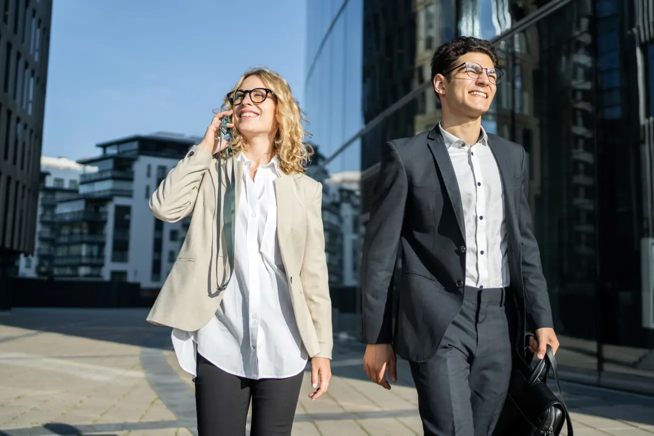 Zwei junge Business-Professionals in moderner urbaner Umgebung, eine Frau mit heller Jacke und Brille telefoniert lächelnd, ein Mann im dunklen Anzug trägt eine Aktentasche, beide gehen selbstbewusst vor einem Glasgebäude entlang.