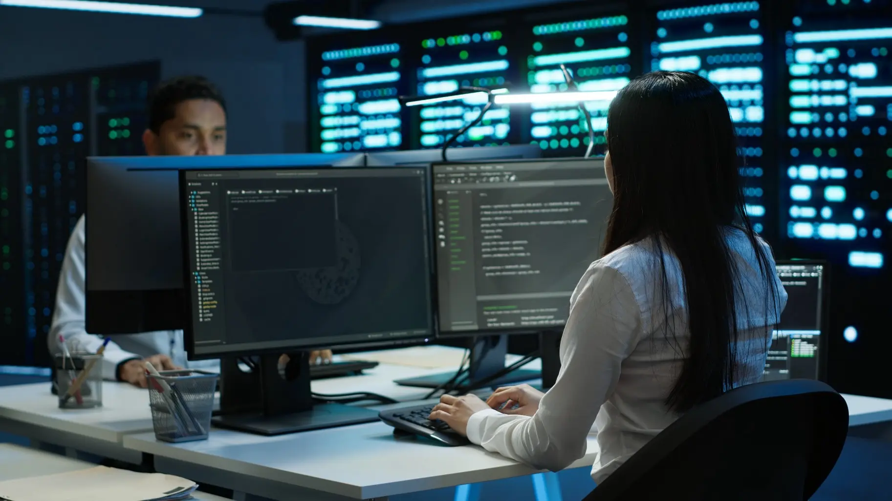 Zwei IT-Fachkräfte arbeiten in einem modernen Rechenzentrum mit mehreren Monitoren, auf denen Programmiercode und Systemdaten zu sehen sind, im Hintergrund leuchten zahlreiche Server mit blauen Statusanzeigen.