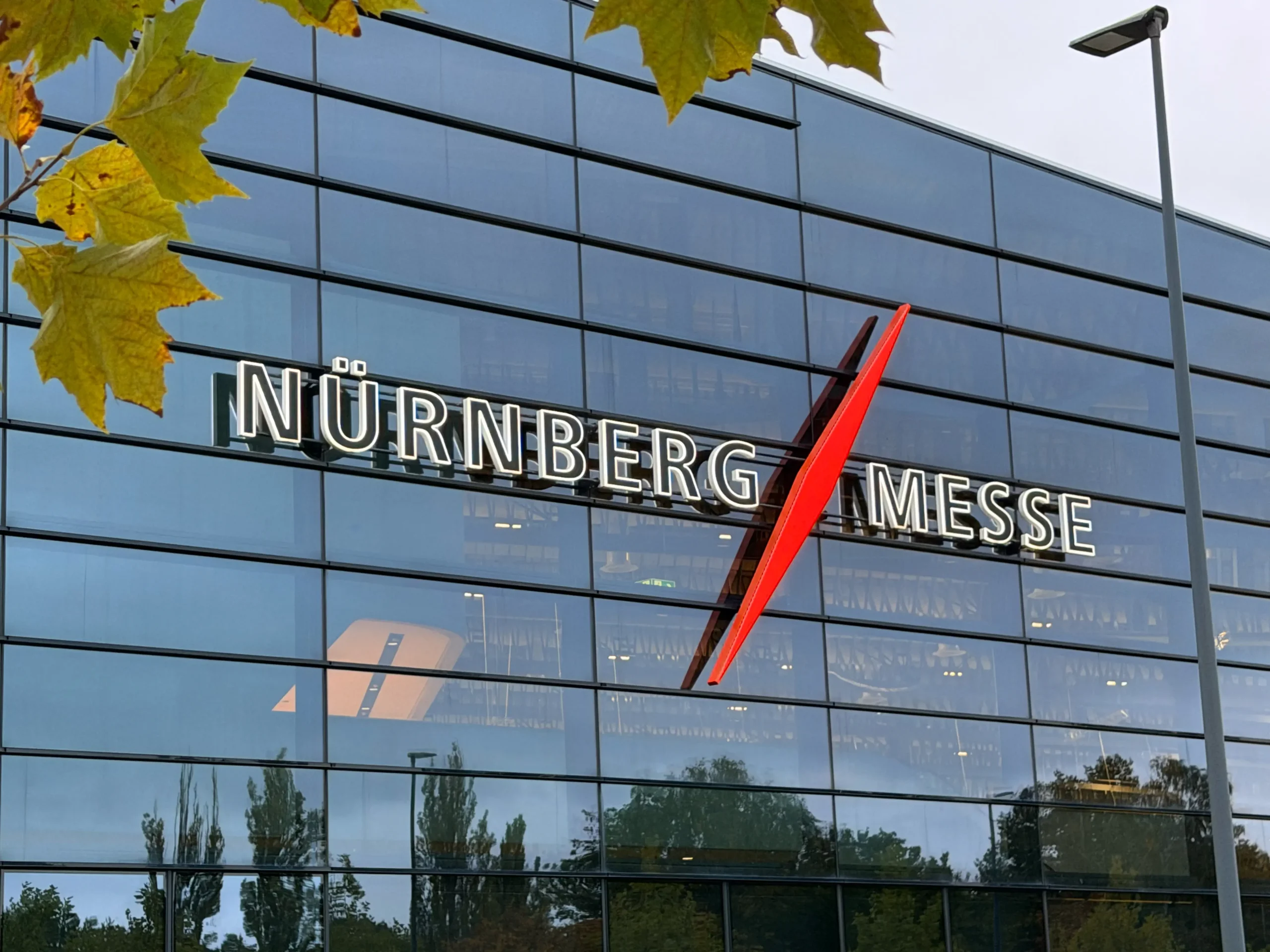 Glasfassade der Nürnberg Messe mit beleuchtetem Schriftzug „NÜRNBERG MESSE“ und rotem diagonalen Akzent, umgeben von herbstlichen Baumblättern und reflektierendem Himmel.