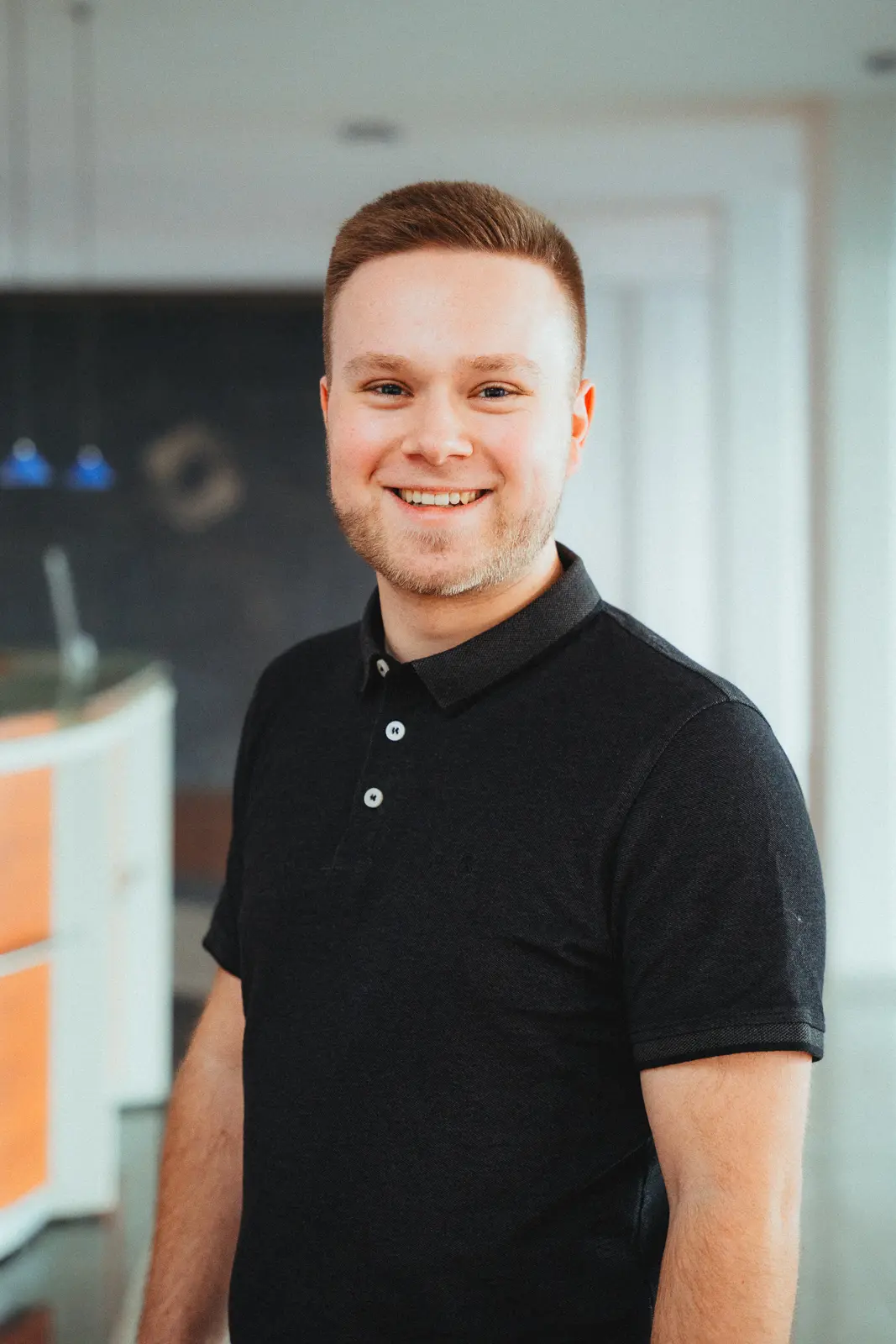 Junger Mann mit kurzem braunem Haar und Bart, lächelnd, trägt ein schwarzes Poloshirt, steht in einem modernen, hellen Büro mit unscharfem Hintergrund.