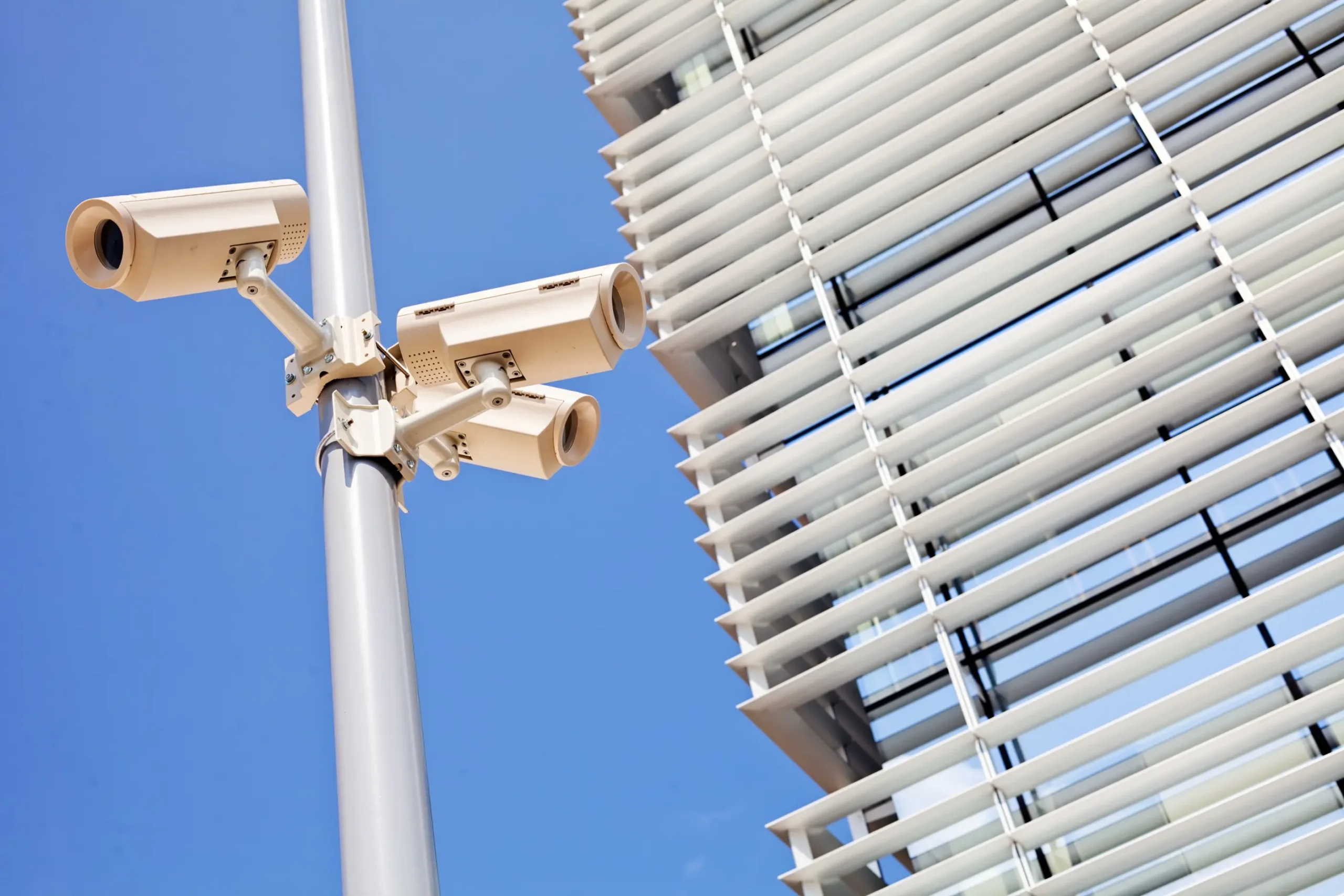 Drei weiße Überwachungskameras an einem grauen Mast vor blauem Himmel, im Hintergrund moderne Gebäudefassade mit horizontalen Lamellen, symbolisiert IT-Security und Netzwerklösungen.